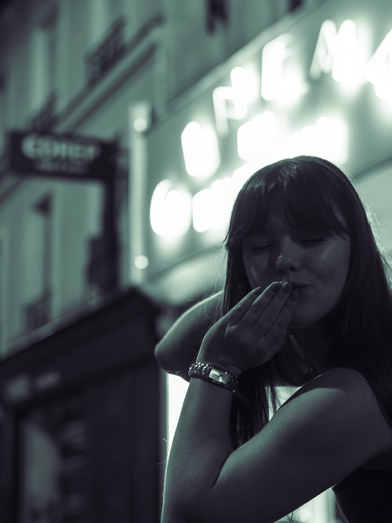 Femme en ville, coiffure longue, s'apprête à faire un geste de baiser. Lumière néon en arrière-plan.