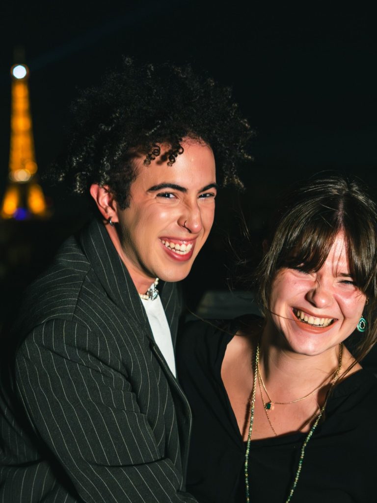 Deux personnes souriantes posent avec la tour Eiffel illuminée en arrière-plan.