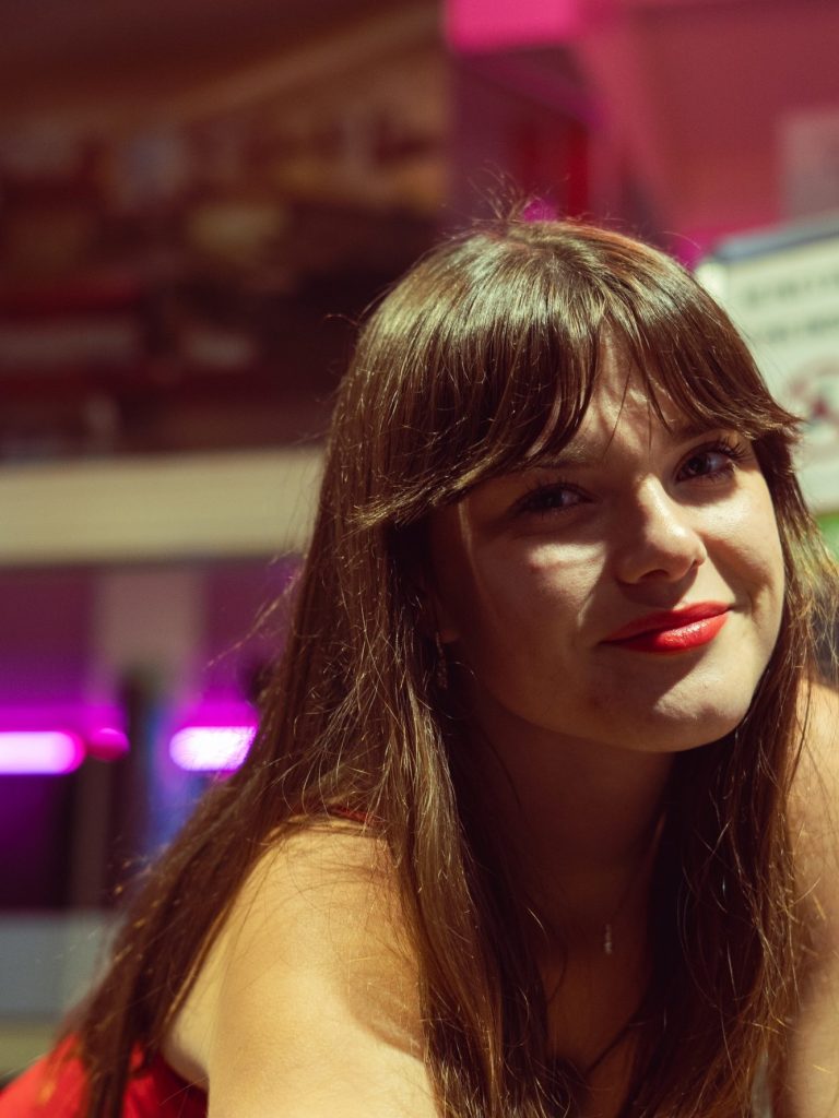 Femme avec des cheveux bruns, sourire, rouge à lèvres et arrière-plan coloré.