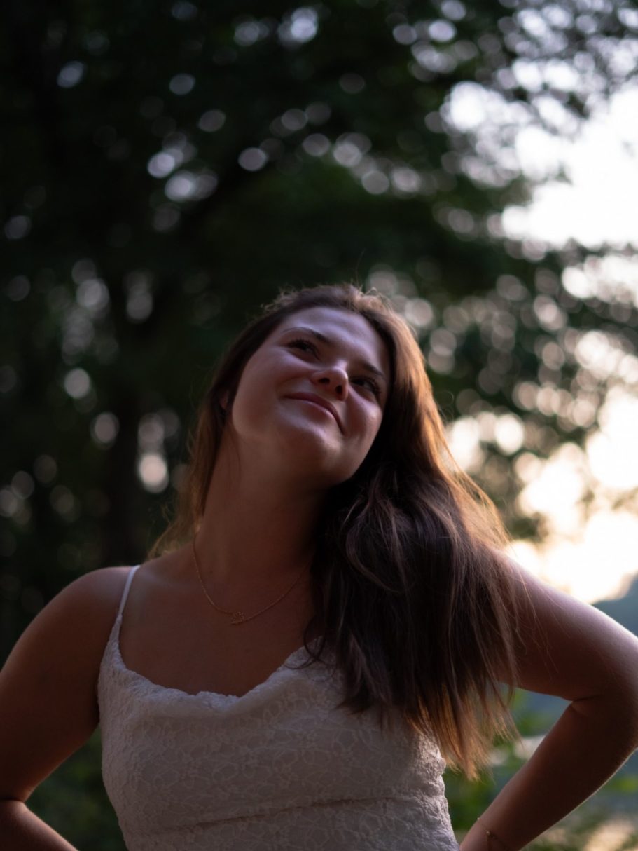 Femme souriante en arrière-plan naturel, cheveux longs, portant un haut léger.