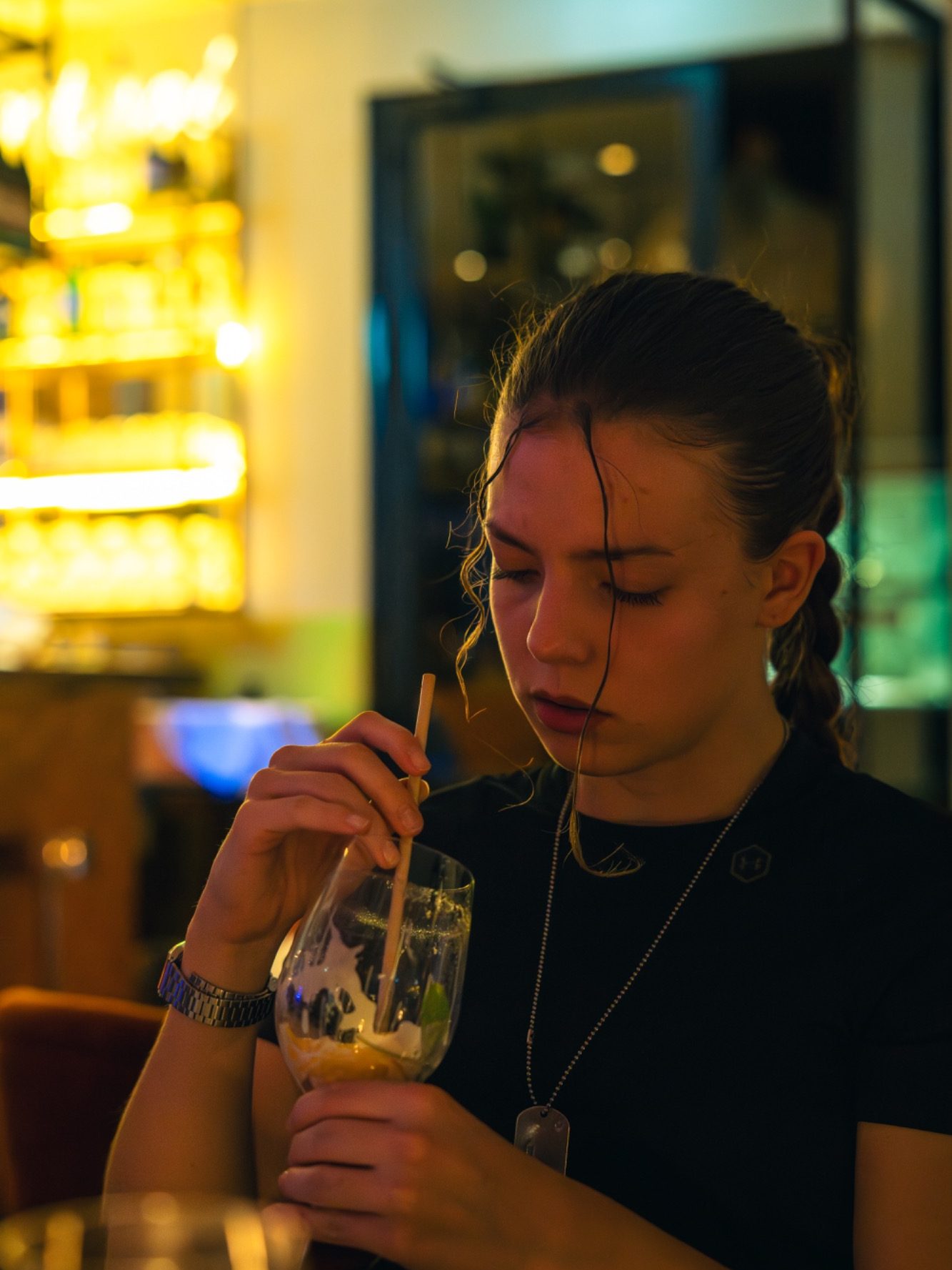 Femme avec une boisson, songeuse, assise dans un bar vibrant et lumineux.