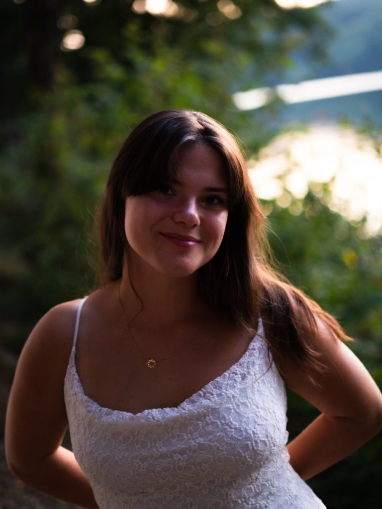 Femme souriante en robe blanche, posant devant un paysage naturel flou.