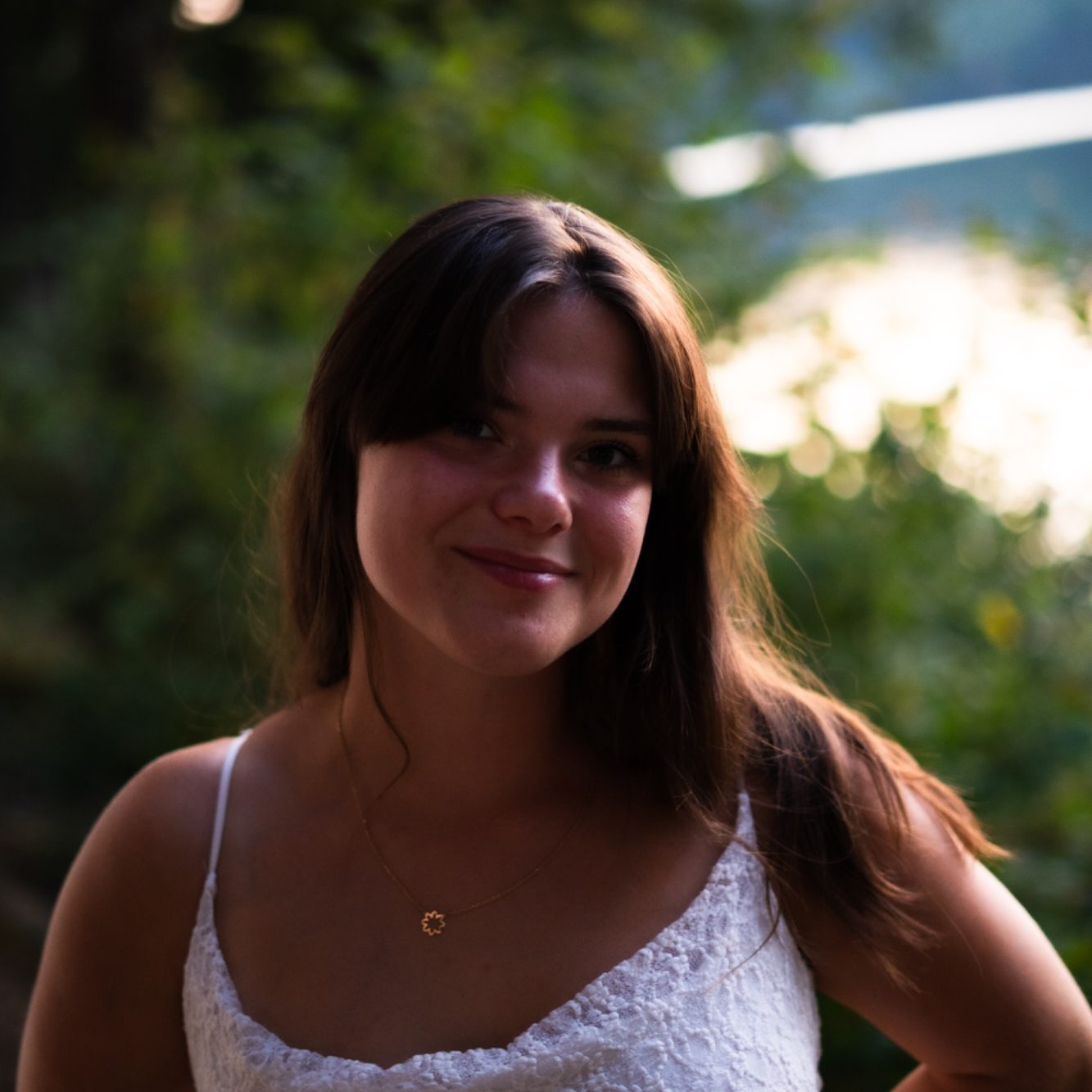 Jeune femme souriante avec des cheveux longs et bruns, vêtue d'un top blanc. Arrière-plan verdoyant.