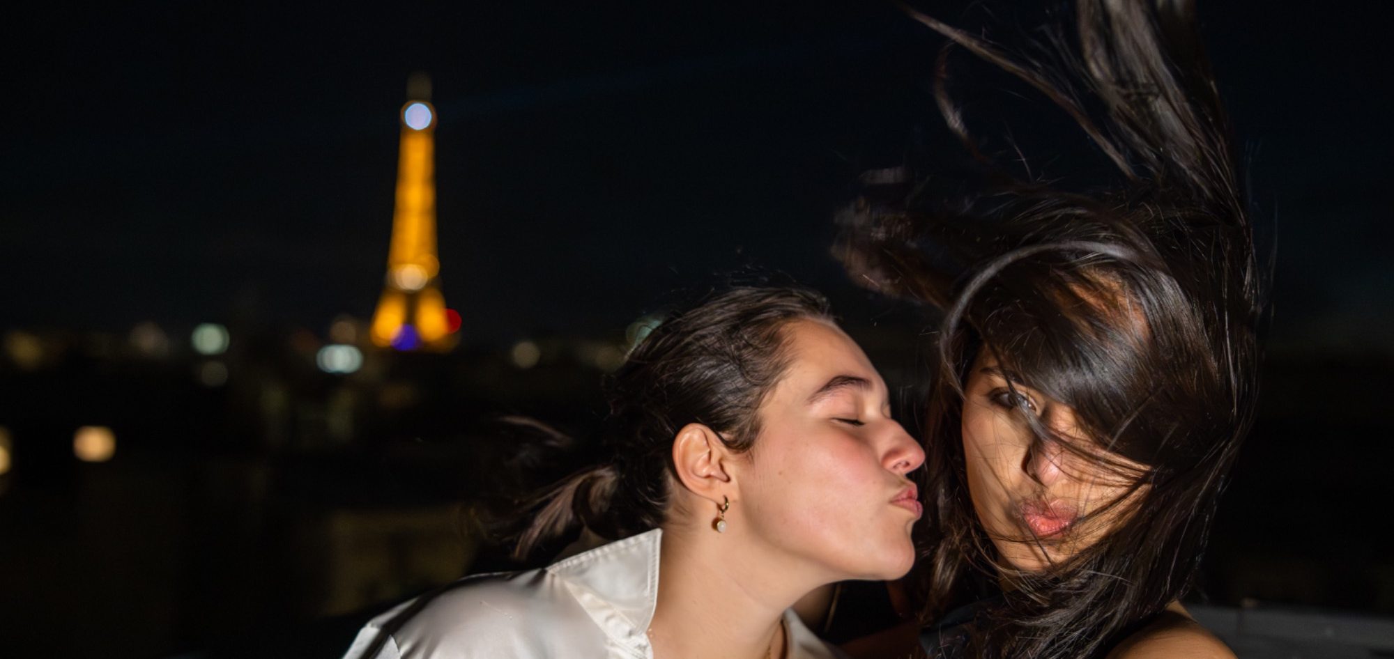 Deux femmes souriantes sur un toit avec la tour Eiffel illuminée en arrière-plan.