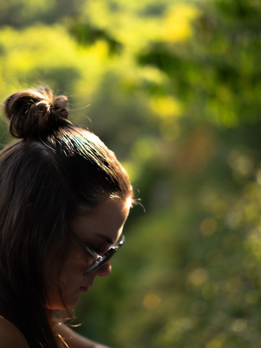 Femme avec des lunettes, cheveux attachés, contemplant un paysage verdoyant.