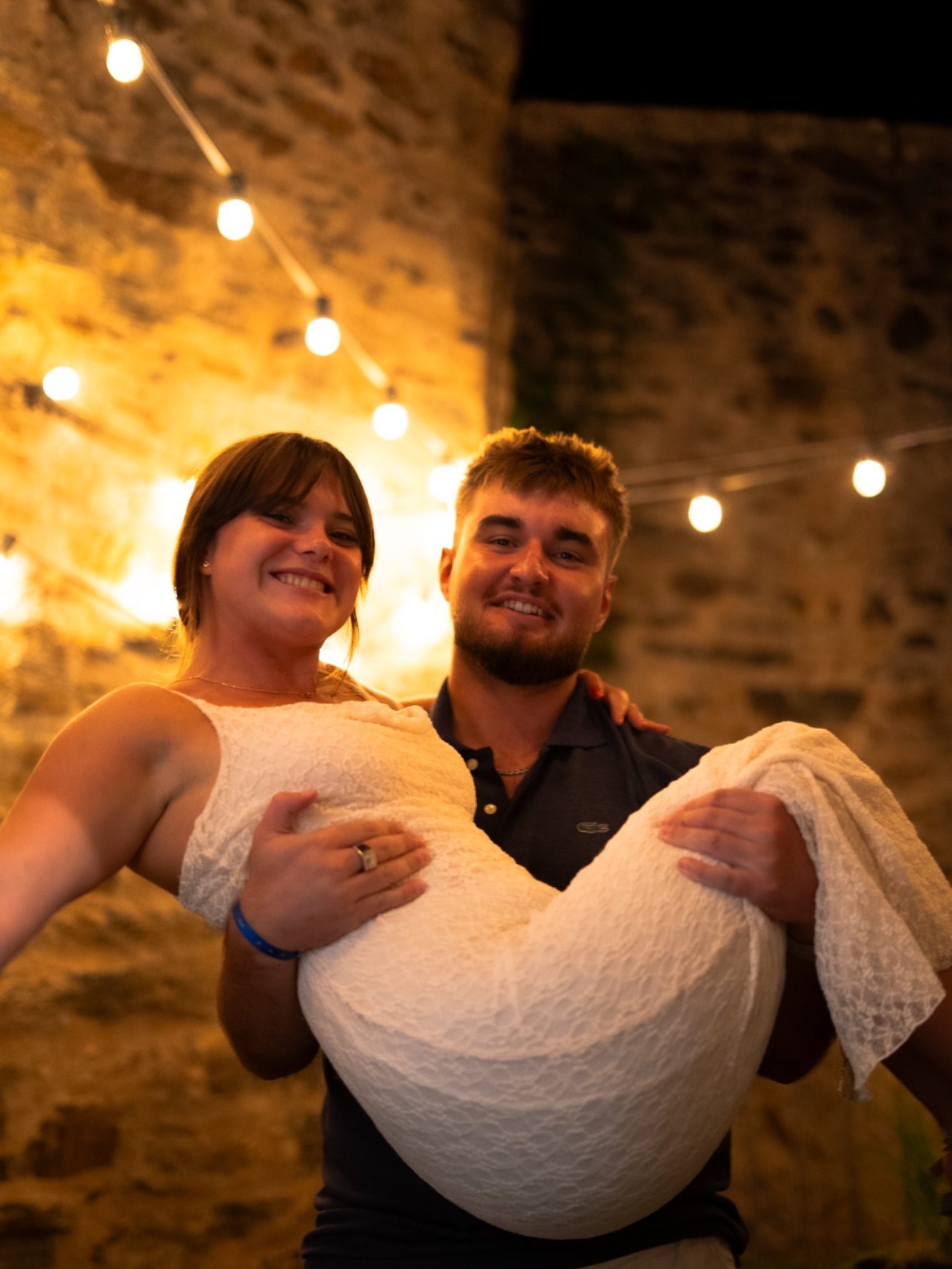 Un homme porte une femme souriante, des guirlandes lumineuses en arrière-plan.
