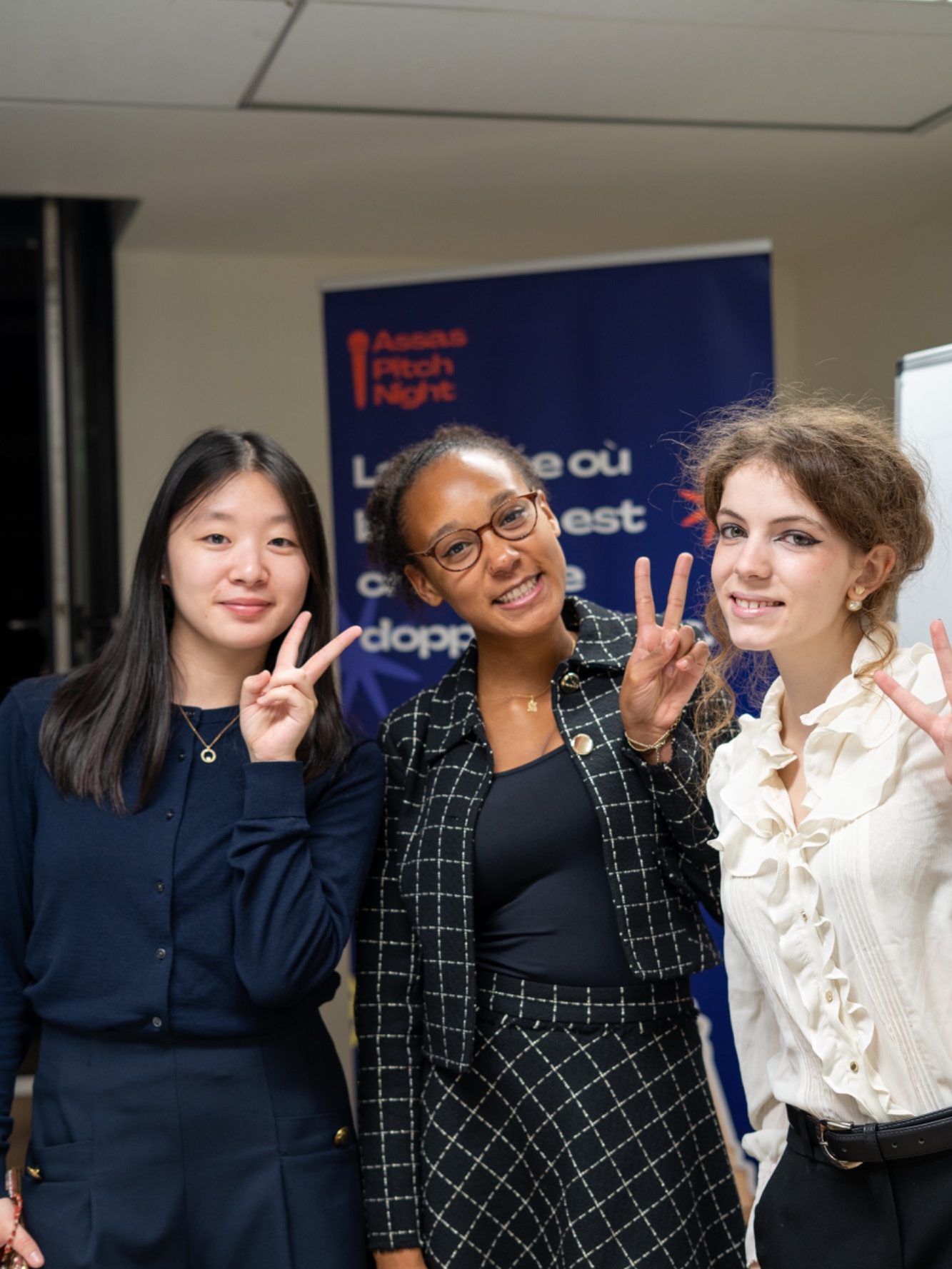 Trois femmes souriantes posent avec des gestes de paix devant une bannière.
