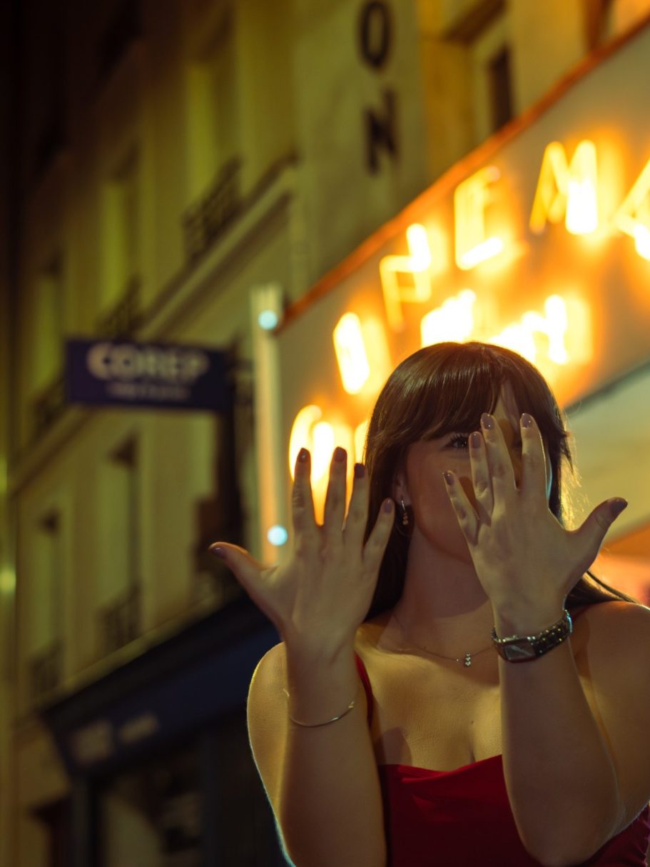 Femme avec les mains levées, devant un cinéma au néon la nuit.