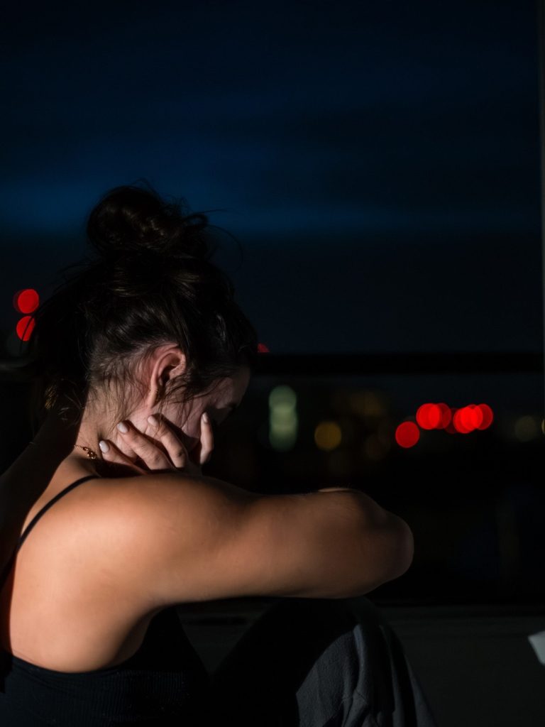 Femme assise, se tenant la tête, dans un environnement sombre avec des lumières floues en arrière-plan.