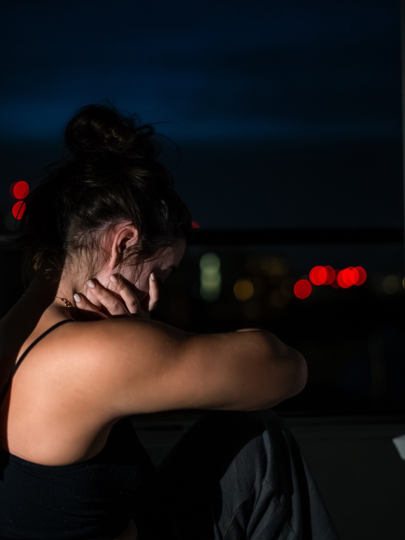 Femme assise dans l'obscurité, la tête inclinée, avec une lumière urbaine en arrière-plan.