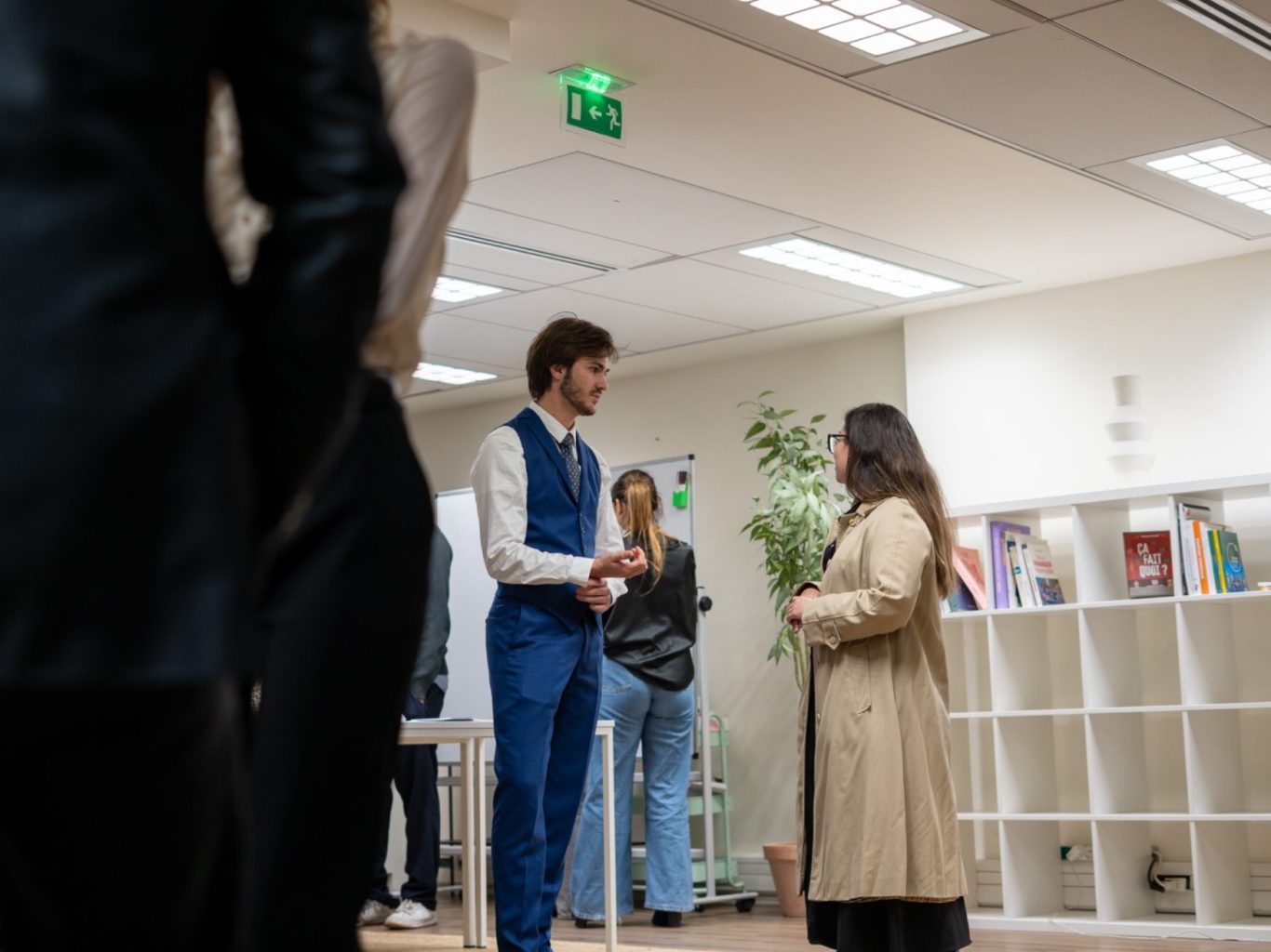 Deux personnes discutent dans un bureau, entourées de livres et d'autres personnes.