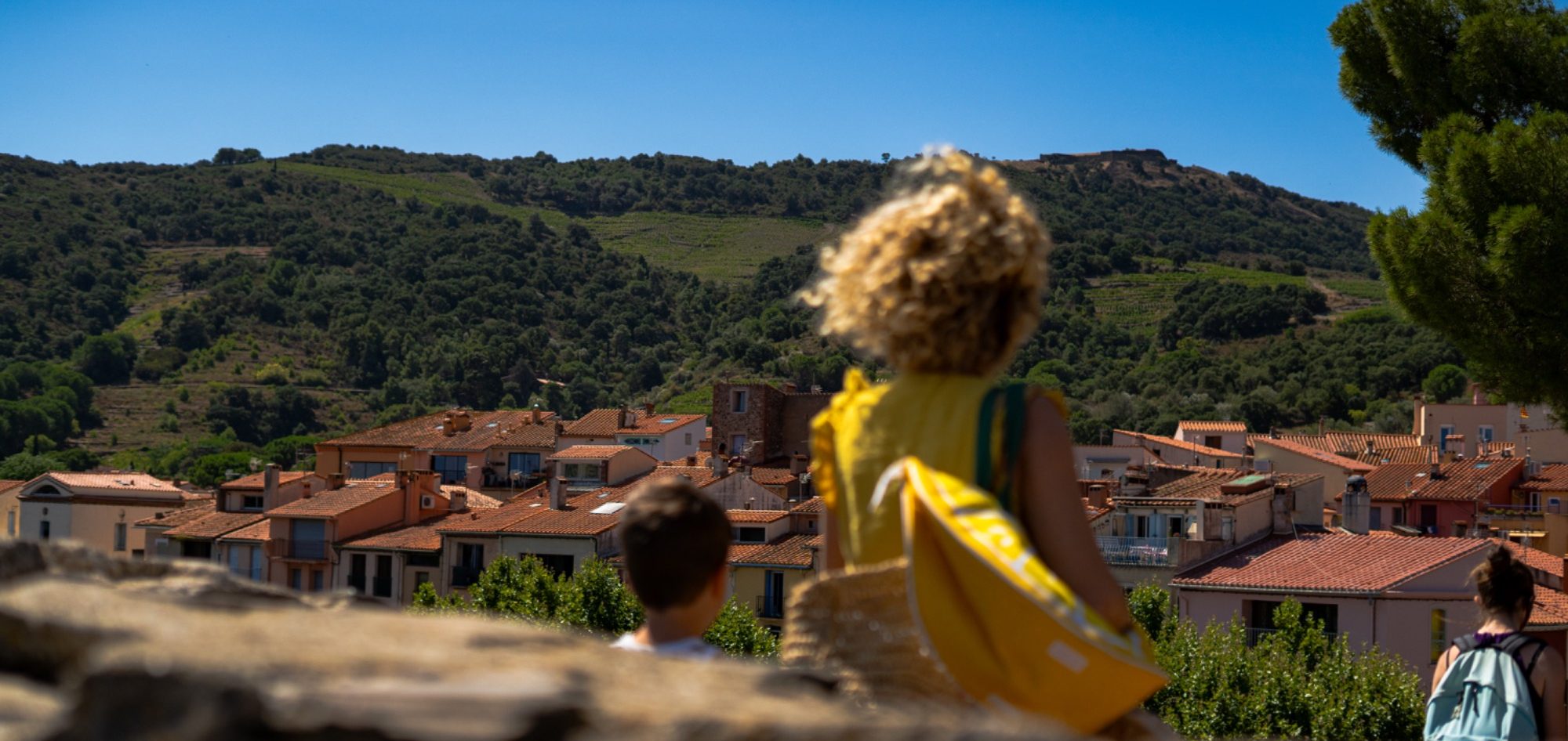 Enfant avec une serviette regarde un village ensoleillé depuis une colline verte.