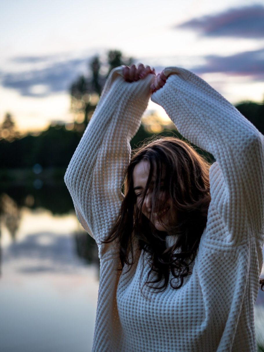 Femme souriante en pull blanc, levant les bras près d'un lac au coucher du soleil.