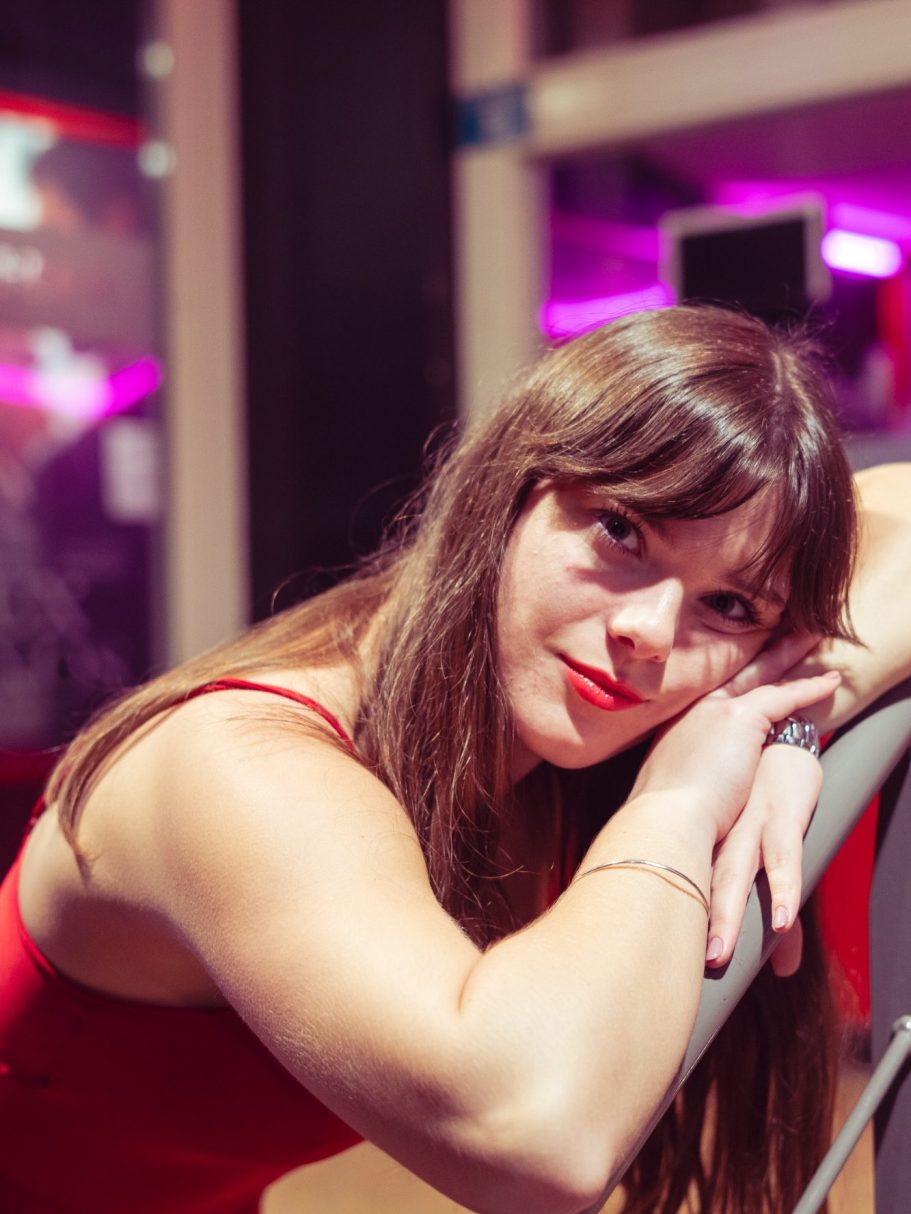 Femme aux longs cheveux bruns, portant une robe rouge, regard pensif.