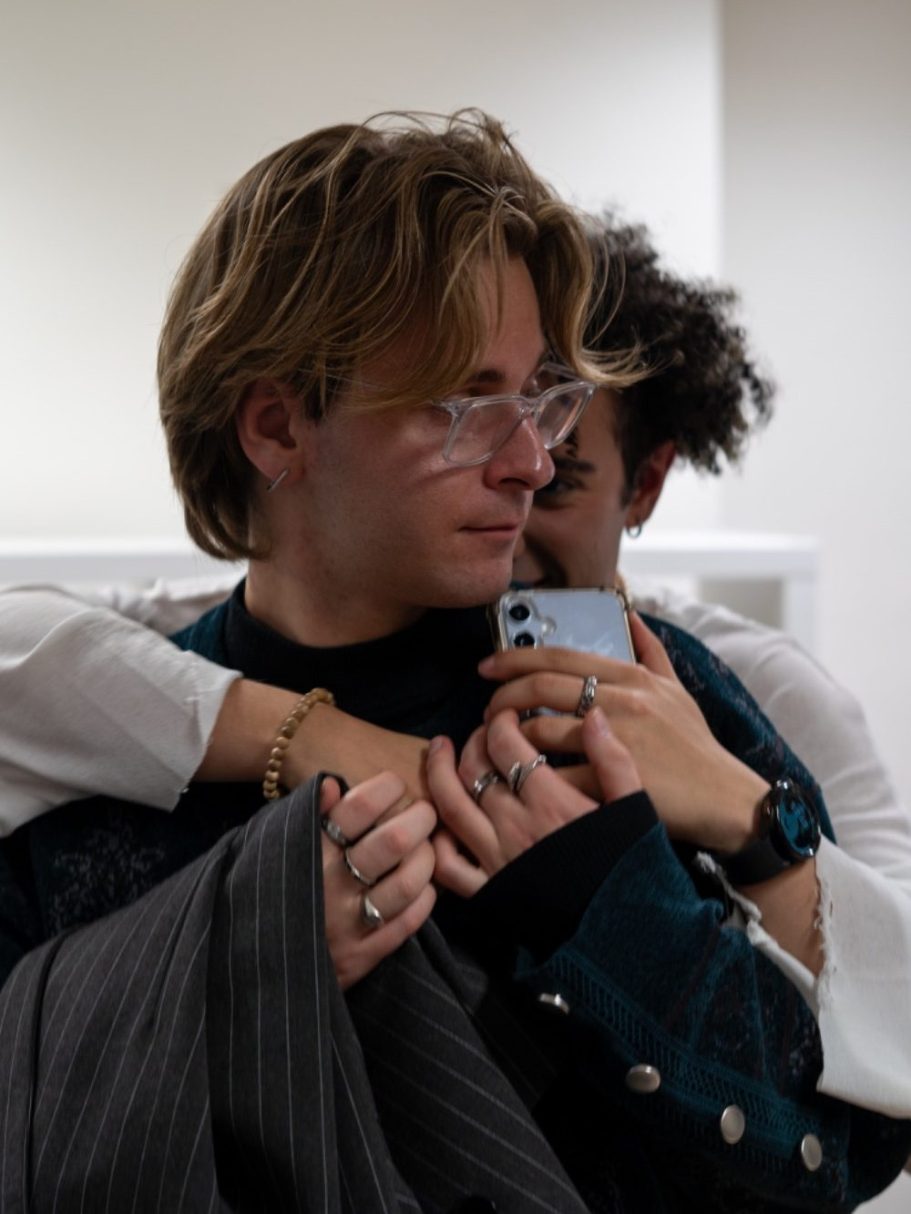 Deux jeunes hommes s'enlacent, l'un tenant un téléphone, dans un intérieur moderne.