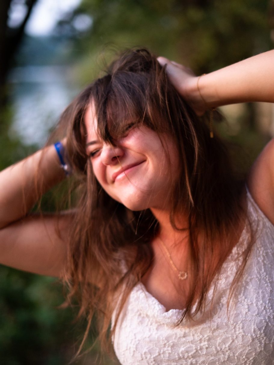 Femme souriante avec les cheveux ébouriffés, posant près d'un lac en arrière-plan.