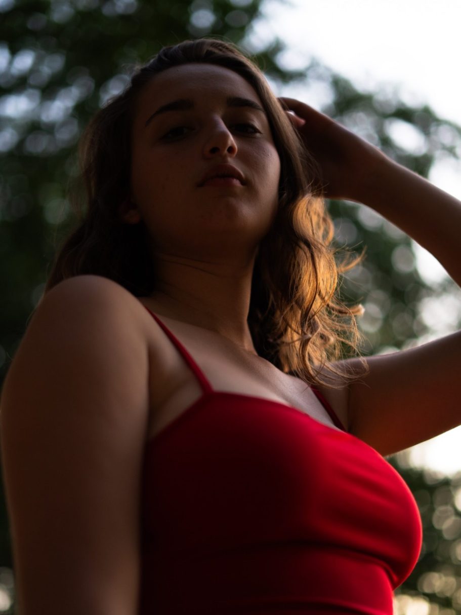 Jeune femme en robe rouge, regard pensif, cheveux au vent, arrière-plan flou.