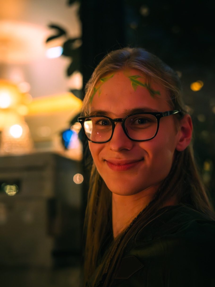 Jeune personne souriante avec des lunettes et des décorations vertes sur le front.