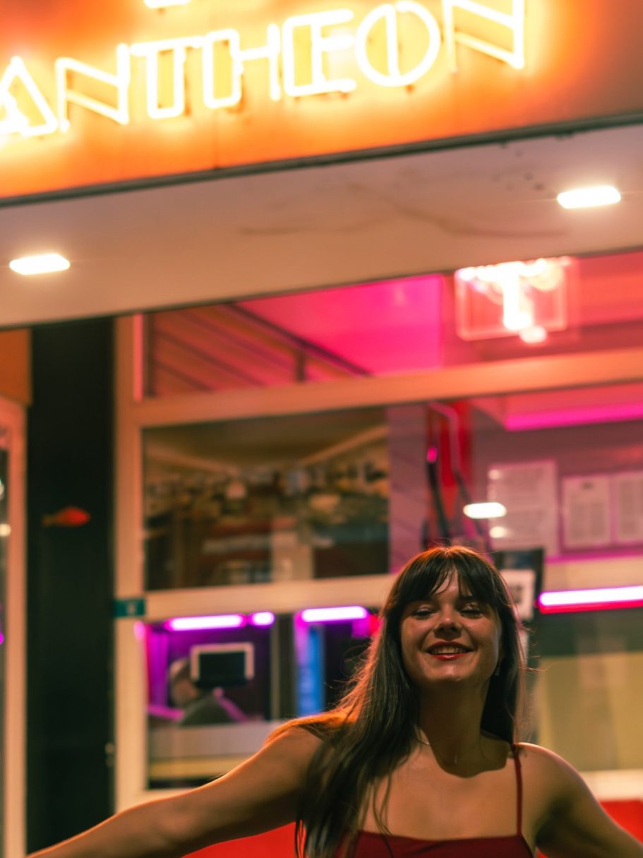 Femme souriante devant un bâtiment avec un néon "PANTHEON" illuminé.