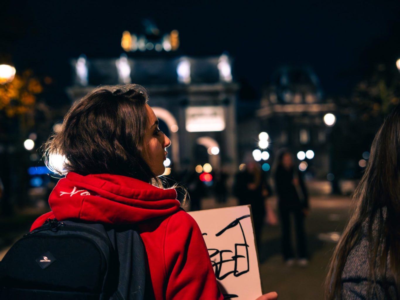 Femme portant un panneau dans une rue illuminée la nuit, monument en arrière-plan.