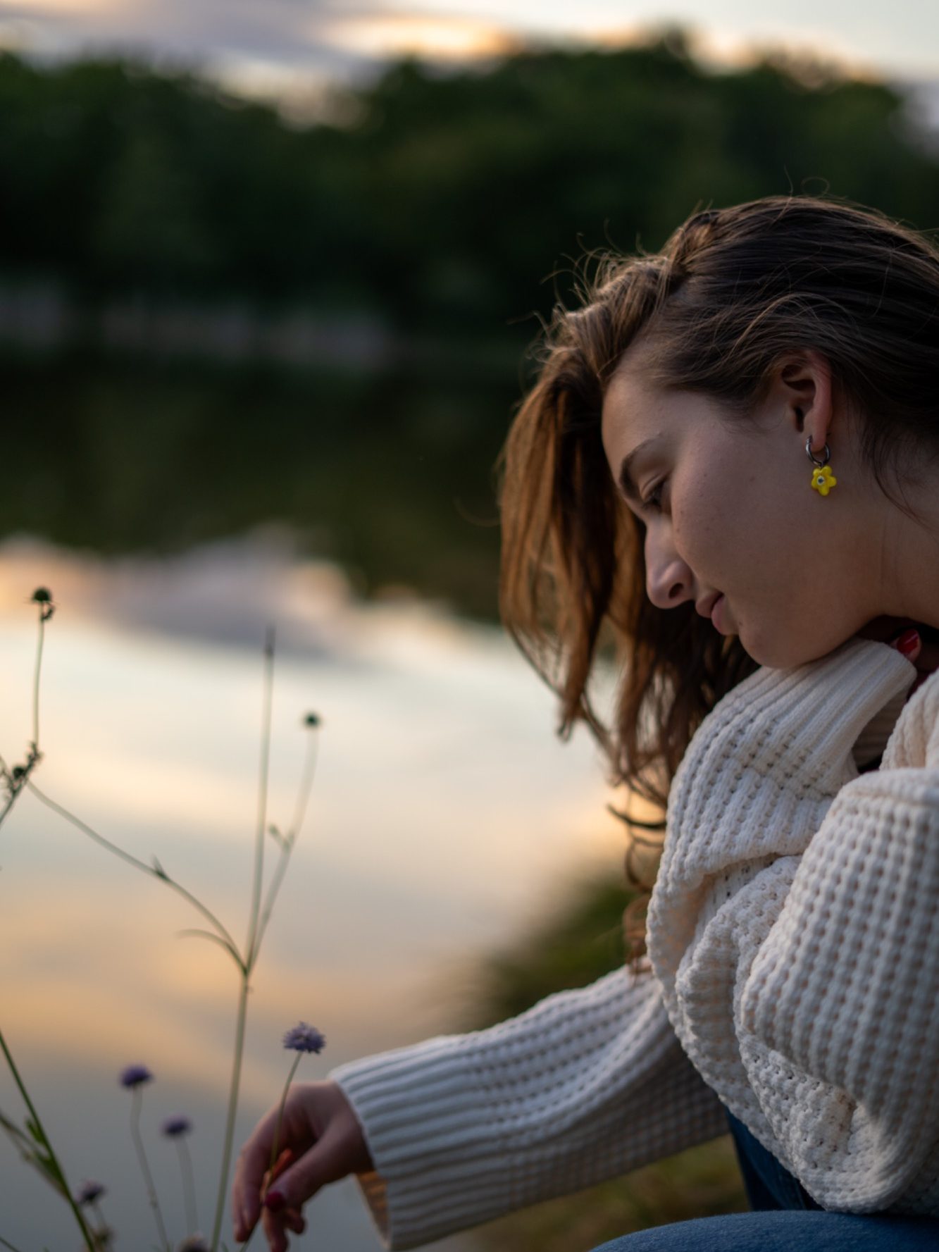 Femme en pull blanc se penchant sur des fleurs au bord d'un lac au coucher du soleil.