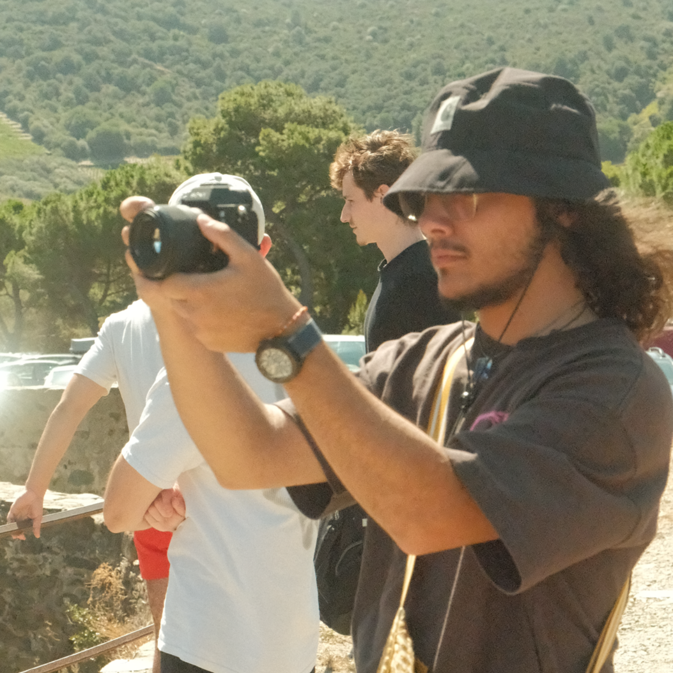 Jeune homme avec un chapeau, prenant des photos, entouré de personnes au loin.