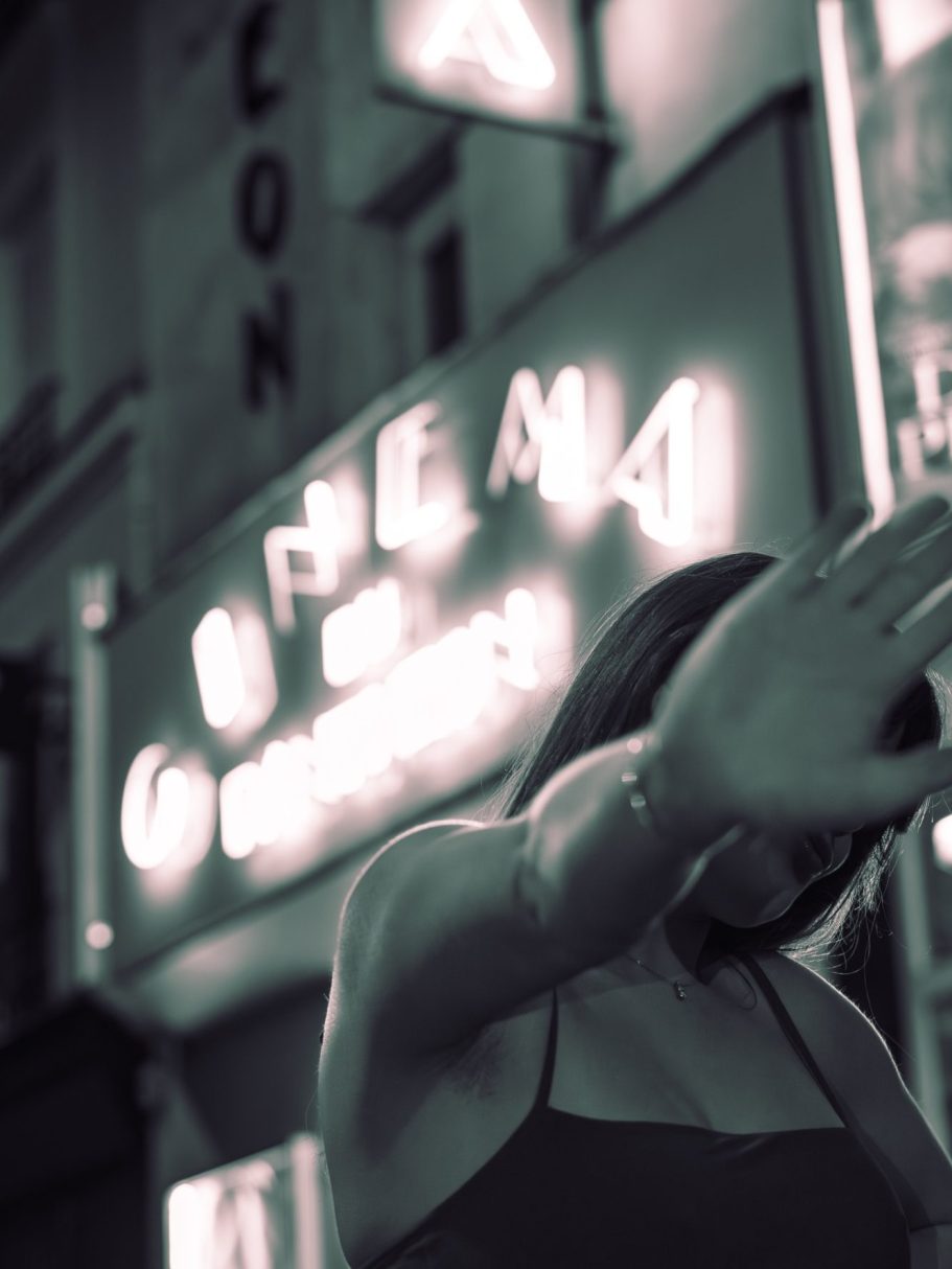 Femme floue en avant-plan, signalant de la main, avec un néon de cinéma en arrière-plan.