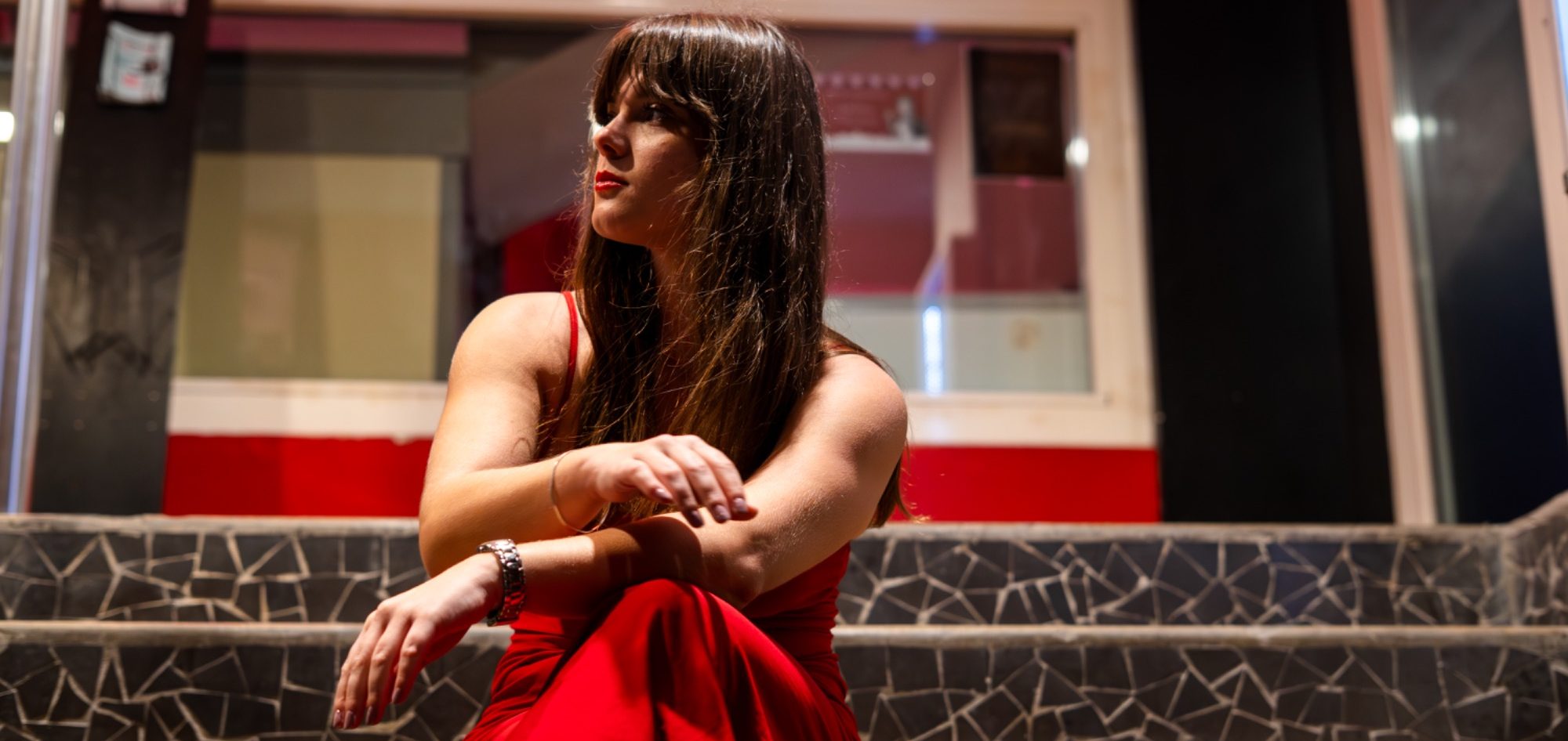 Femme assise sur les marches, portant une robe rouge, regard pensif vers l'extérieur.