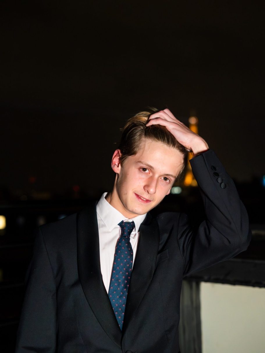 Jeune homme en costume, posant avec la tour Eiffel en arrière-plan nocturne.