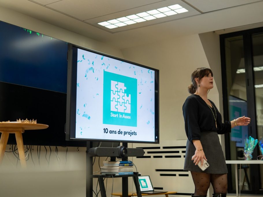 Une présentatrice debout devant un écran, parlant à un public dans un espace lumineux.