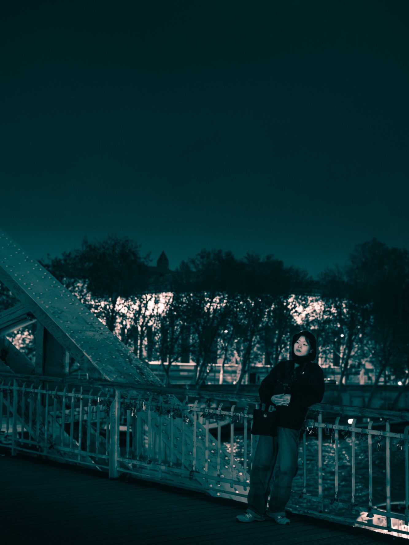Femme debout près d'un pont illuminé, nuit sombre avec des arbres en arrière-plan.