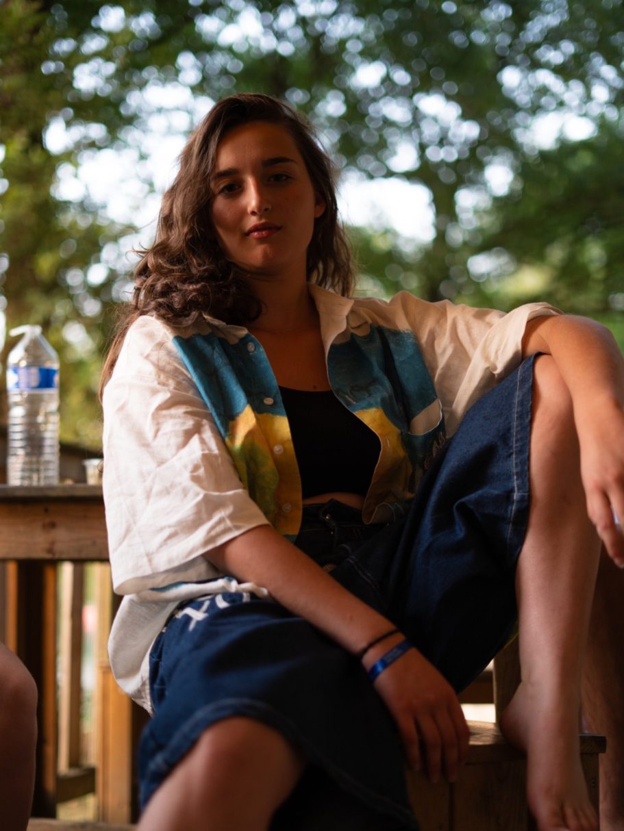 Une jeune femme assise sur une terrasse, portant une chemise colorée et un pantalon large.